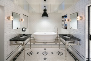 The en-suite master bathroom has a free standing tub by the Water Monoply , dual Urban Archaeology vanities with Gringio Carnico marble tops, a shower, white tiled walls and a white and black patterned tile floor.