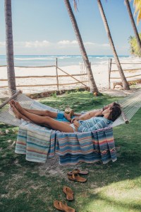 A couple reclines in textured Faherty beach clothing on a hammock spread with folk blankets with a palm landscape in the background.