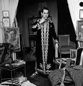American accessories designer Kenneth Jay Lane wears an embroidered caftan with matching slippers.  He stands, smoking a cigarette, in a room with chairs, a zebra print rug, and various paintings dispalyed on easels.
