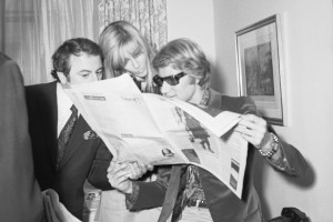 Yves Saint Laurent reading Women's Wear Daily with Pierre Berge and model Betty Catroux after arriving in New York prior to attending a party given by Andy Warhol at the downtown Factory location.