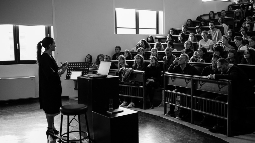 A black and white image of CFDA's Sara Kozlowski teaching a class at Polimoda with a stadium-style seating at side profile.