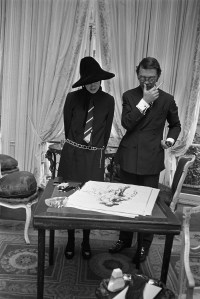 WWD illustrator Kenneth Paul Block stands at a table with Gloria Vanderbilt Cooper, showing her his sketch of her.