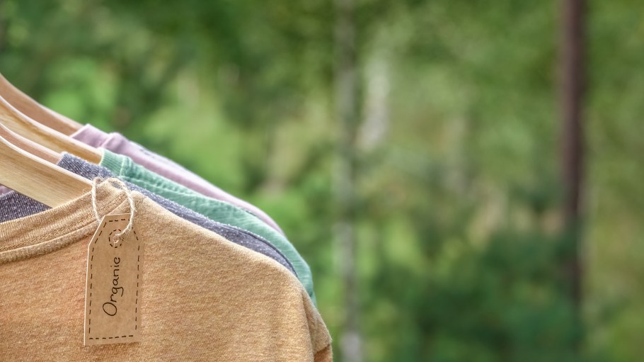 Organic clothes. Natural colored t-shirts hanging on wooden hangers in a row. Eco textile tag. Green forest, nature in background.