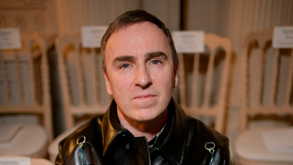 Raf Simons sits on a char in a black leather coat.