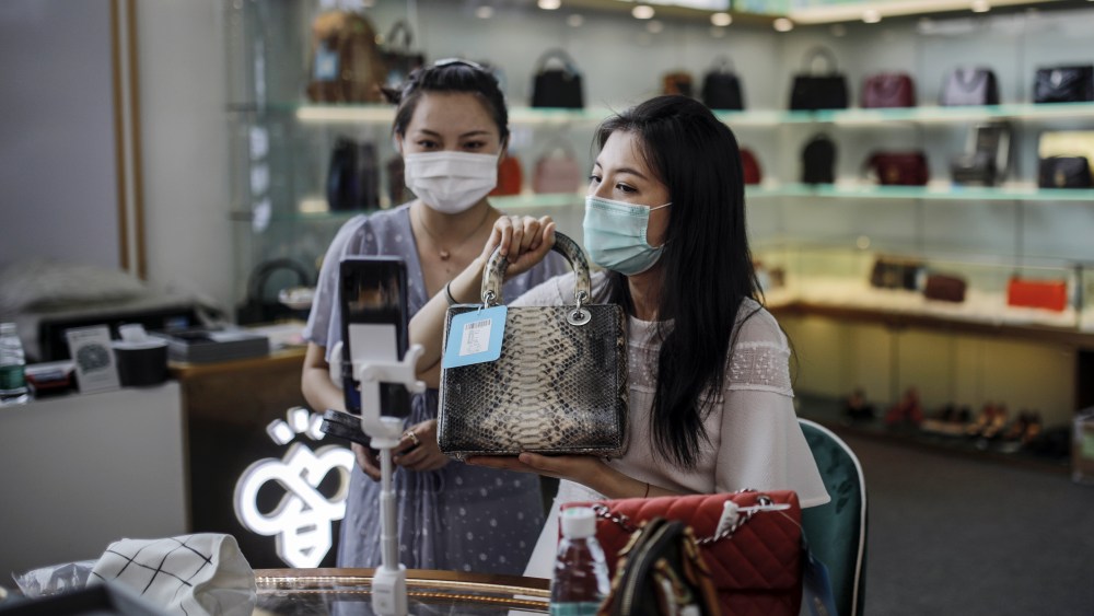 A saleswoman wearing protective face mask holds a handbag as she uses mobile phone to do live stream for consumers in a luxury good store in a shopping mall following more new COVID-19 cases being found recently in Beijing, China, 21 June 2020. Beijing reported new 22 confirmed COVID-19 cases on 20 June.Daily life amid coronavirus pandemic in Beijing, China - 21 Jun 2020