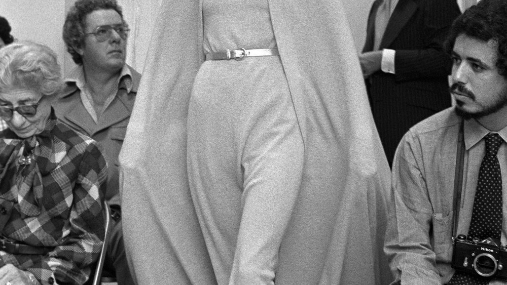 Anjelica Huston models a belted knit gown and matching hooded cape on the runway of the Halston resort 1972 RTW collection.