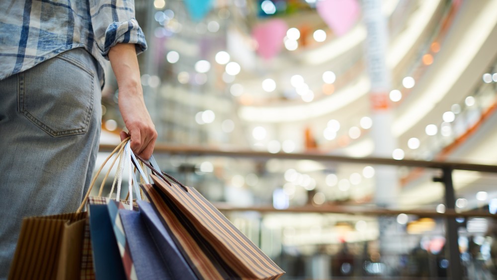 Close-up of unrecognizable woman in casual clothing standing in spacious shopping mall with lights and holding many paper bags