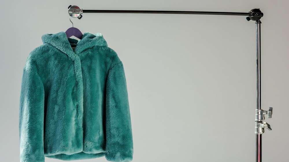 fluffy green faux fur coat hanging on rack at grey background with sunbeams