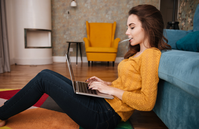 Portrait of a young woman doing shopping online and holding credit card in her mouth while sitting in the living room at home