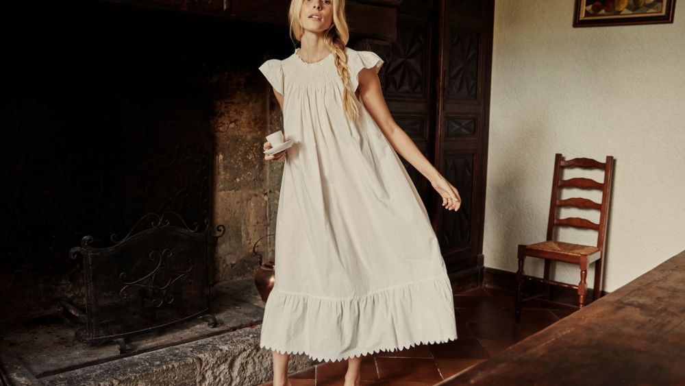 A model in an off-white Dôen nightgown holding a teacup and tilted in a warm-toned room with fireplace.