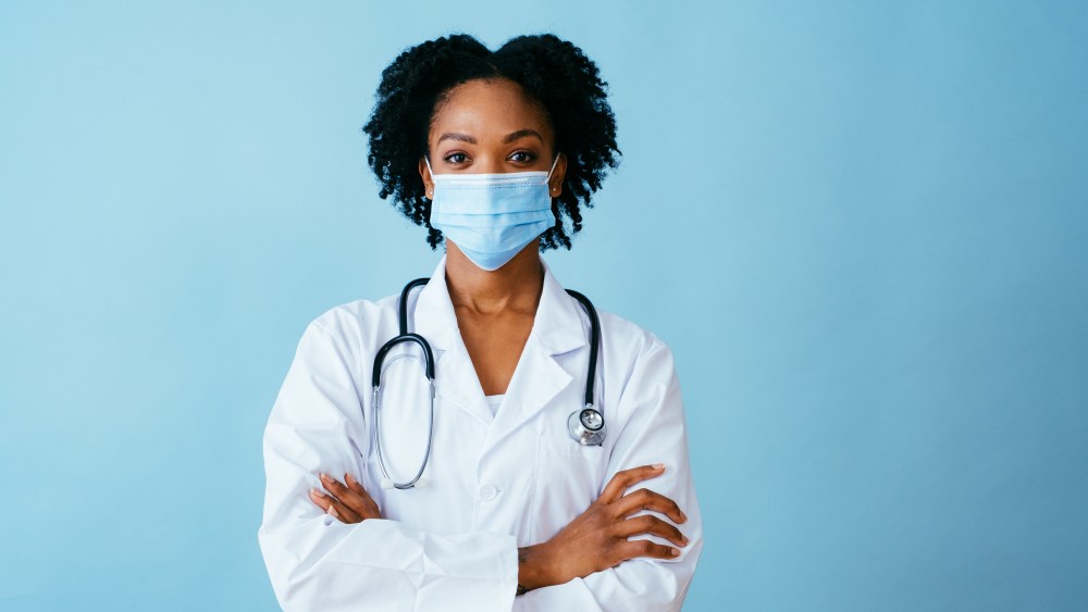 Portrait of a doctor in lab coat with face mask and arms crossed, isolated on blue background