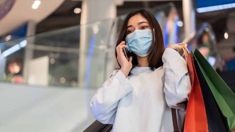 young woman talking with mobile phone in shopping mall and wearing face mask protective for spreading of coronavirus(covid-19) pandemic, new normal concept