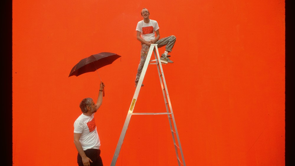 Keith Haring sits on a ladder, in front of the bright orange undercoat on which he will shortly paint his mural Crack is Wack. New York Park Commissioner Stern holds the umbrella. (Photo by Owen Franken/Corbis via Getty Images)