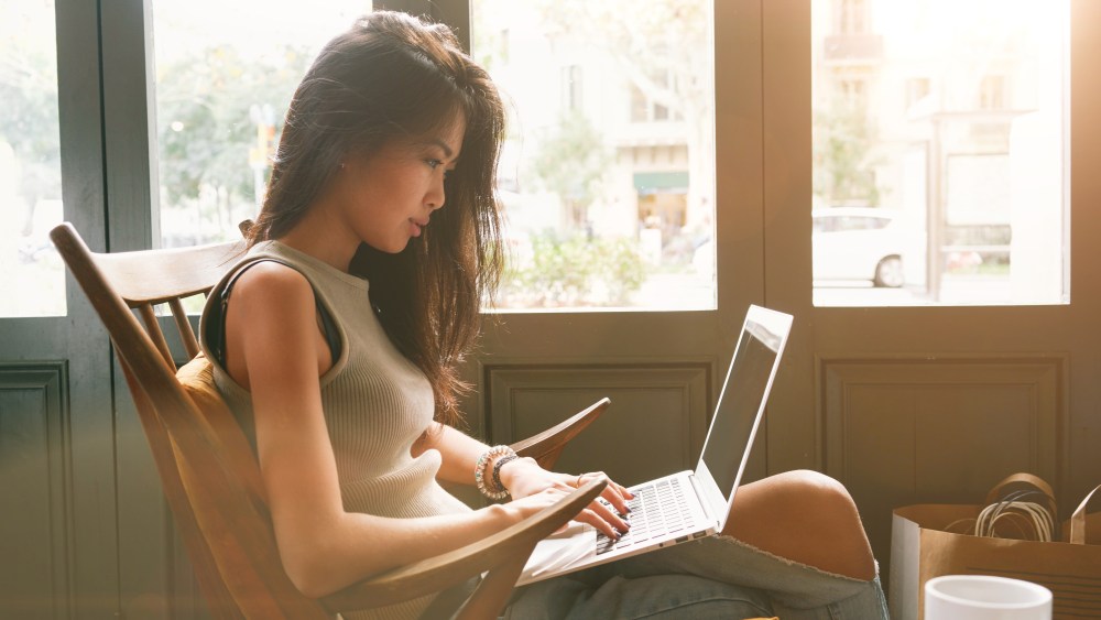 Attractive asian female is typing messages on a laptop while sitting against the wide windows of a coffee shop. a hipster girl is working on a portable computer in a cafe.
