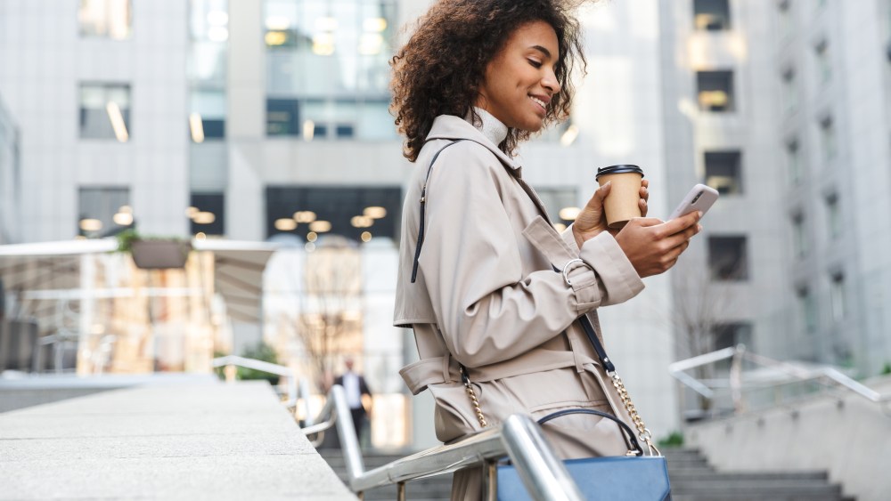 Smiling attractive young african woman wearing autumn coat walking outdoors, using mobile phone
