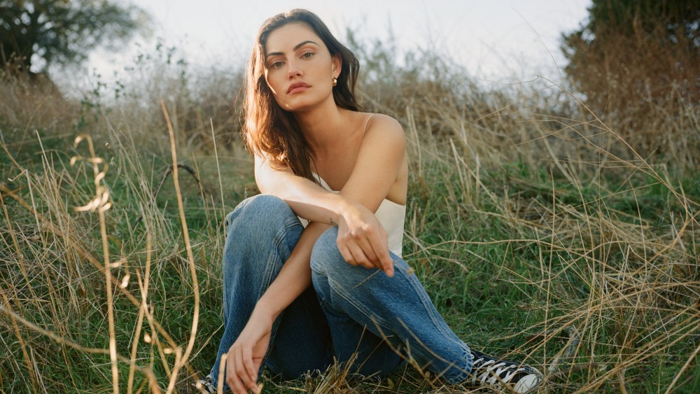 A brunette model sports jeans and tank for Reformation, perched amid a grassy field crossing her arms and gazing softly onward.
