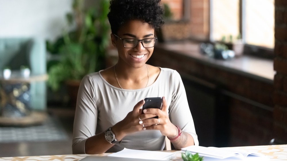 Cheerful african student black woman sitting at table studying using laptop reading a book, take a break holding mobile phone surfing internet received message from friend chatting about weekend plans