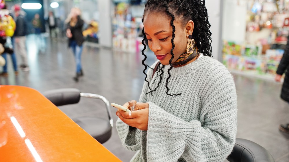 African woman bloger posed in sweater and jeans posed at mall with her mobile phone.