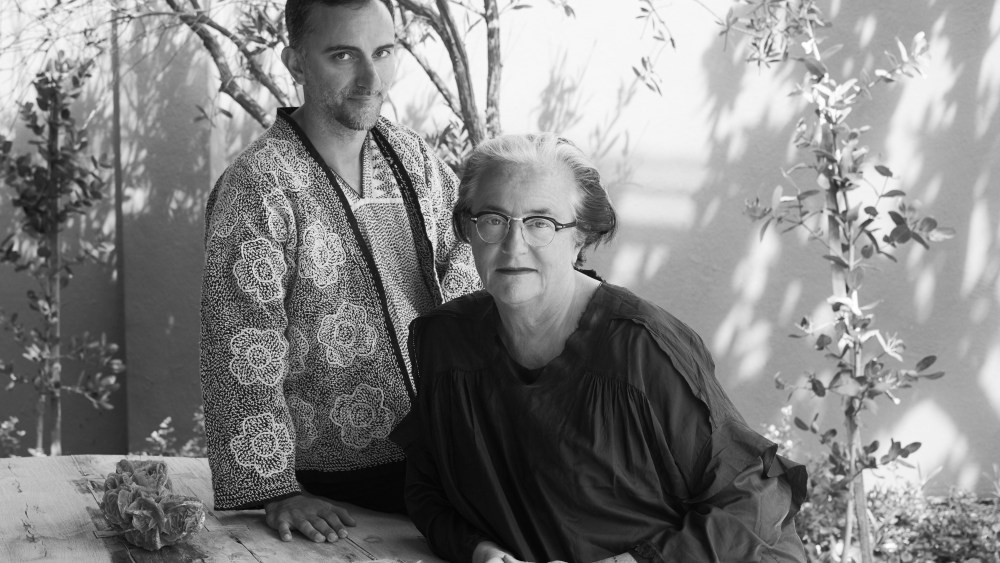 Lidewij Edelkoort and Philip Fimmano pose for black and white photo sitting at a table with tree branches and leaves behind them.