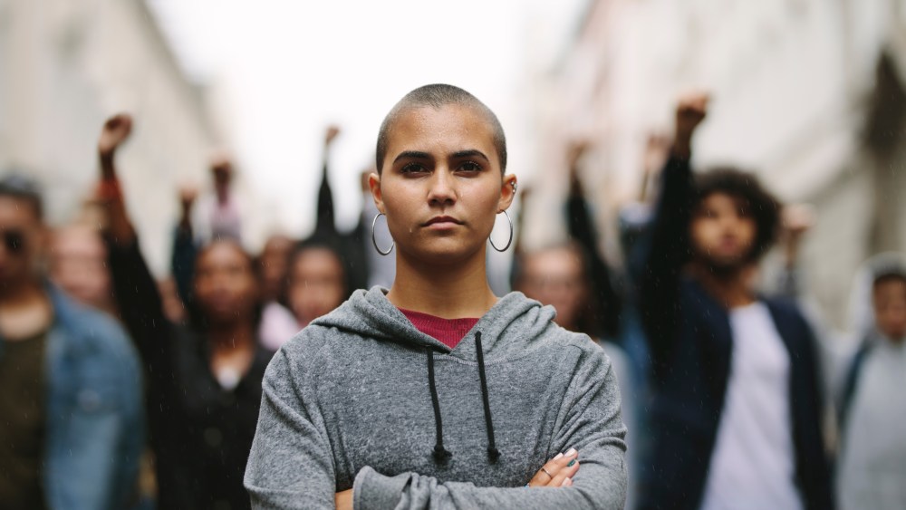 Young woman protestor standing with her arms crossed and group of people in background on the city street. Activists demonstrating in the city.