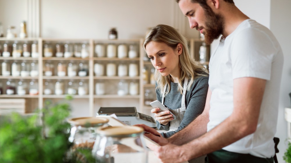 Two shop assistants standing at the counter in zero waste shop, checking and ordering stock.