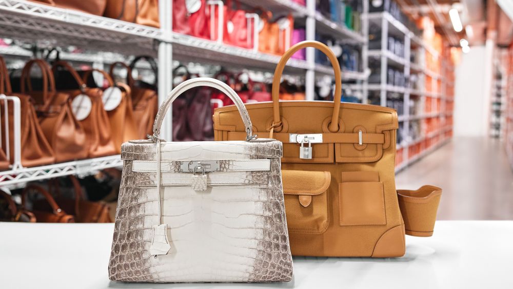 Fashionphile's cage inventory room in background with an array of product and two Birkin bags at front foreground in neutral tones.