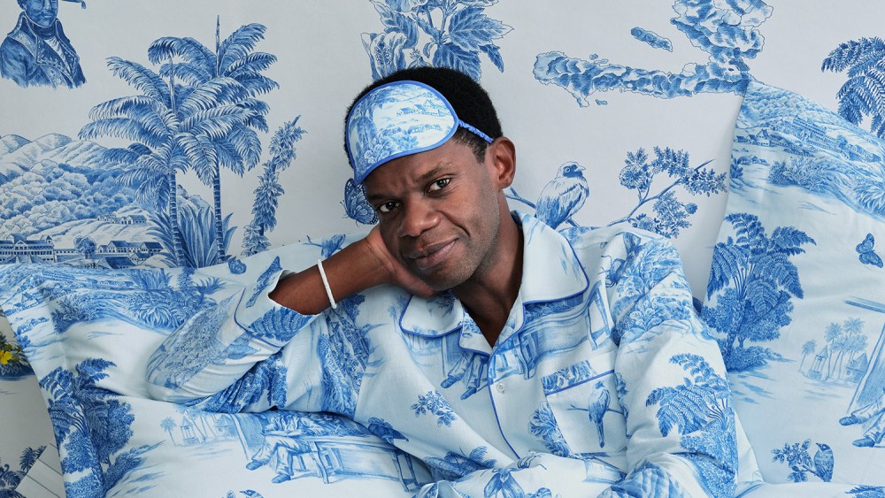 Designer Victor Glemaud sits in a bed with duvet cover and pillowcases in the pattern of his Toussaint Toile that launched as part of his first home goods line with Schumacher. He is also wearing pajamas of the same print and the bed is in front of a wall with wallpaper in the same print.