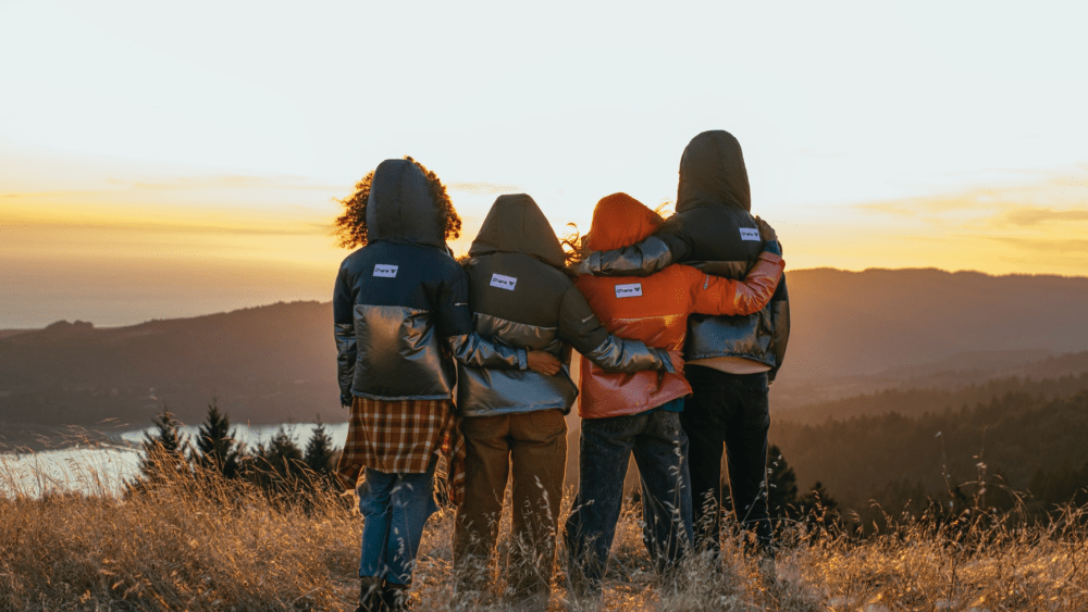 Friends stand shoulder to shoulder watching sunset over mountain wearing recycled outerwear in a range of colors.