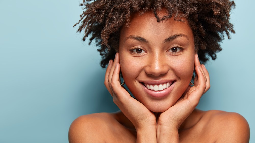 Close up portrait of relaxed black woman has gentle skin after taking shower, satisfied with new lotion, has no makeup, smiles tenderly, shows perfect teeth, stands shirtless against blue background