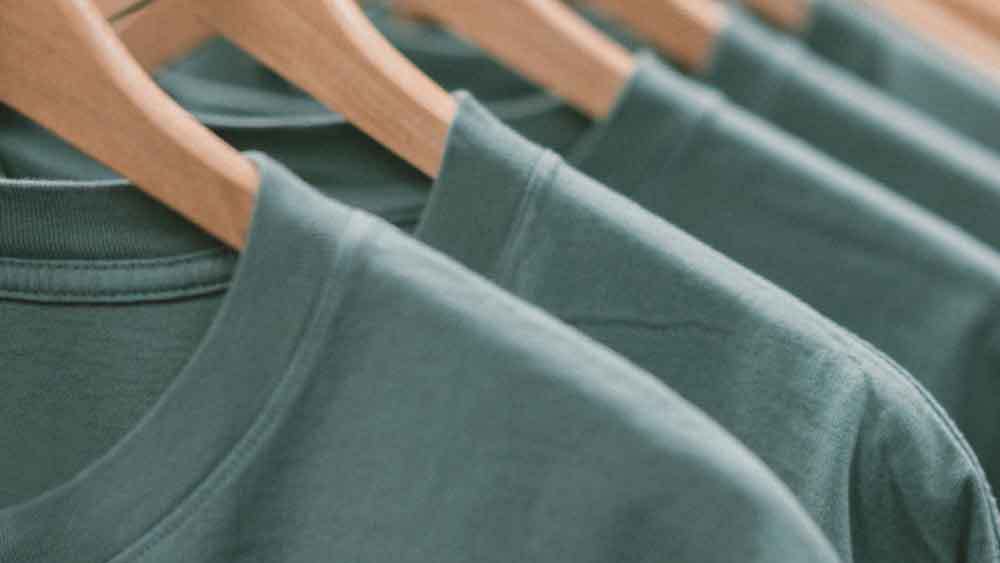 A close-up of sage green cotton t-shirts on a garment rack with wooden hangers.