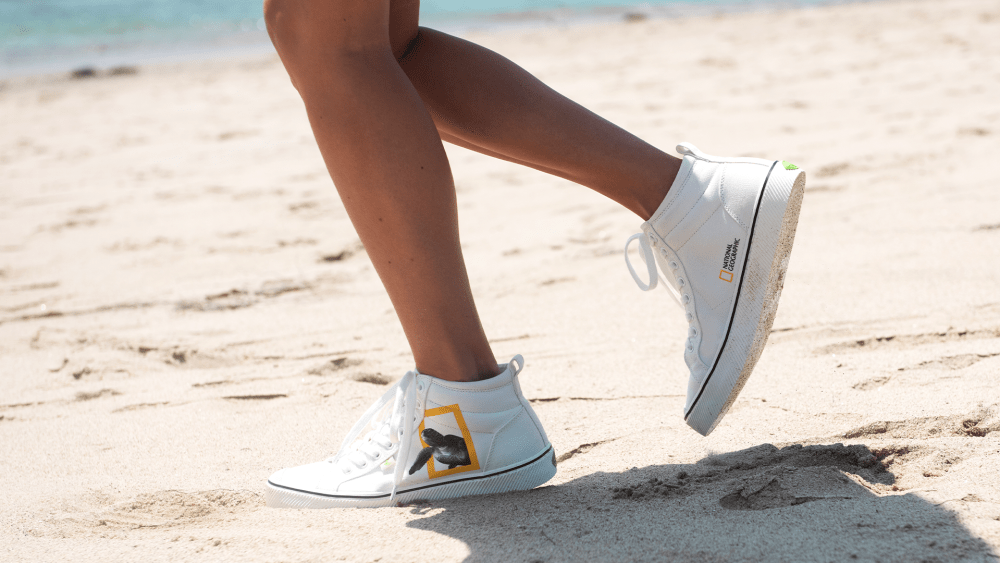 A model strides with a white high-top Cariuma sneaker on the beach. The shoe has a Green Turtle decal on it.