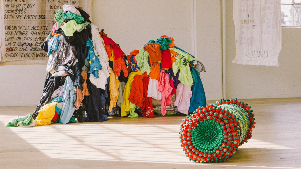 A web of colorful clothing in background of tan white-box exhibit space. At right is a wheel of textured chips of material showcasing sustainable innovation.