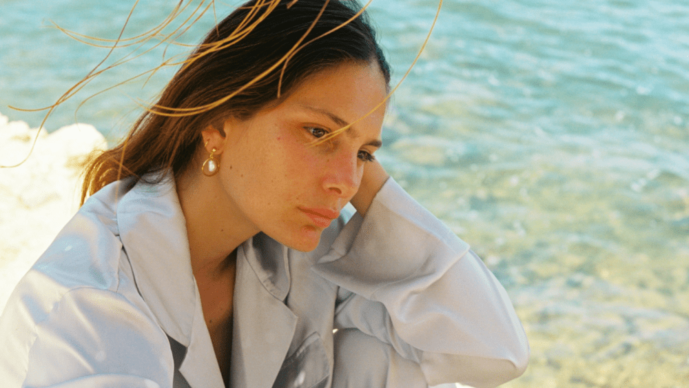 A woman tucks hair begin her ear on the beach, reveling a lab-grown diamond earring.