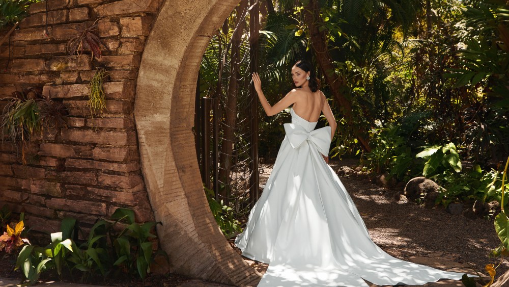 A piece from Nadia Manjarrez Studio Bridal shot by Raul Tovar, features a woman in wedding dress with her back to the camera and a large bow at her back. She is standing next to a stone arch in a garden filled with tropical plants.