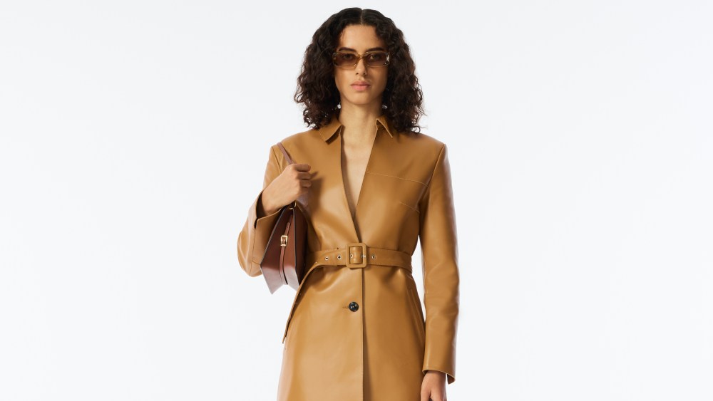 A look from Wandler spring 2023 shows a person with curly hair wearing a tan leather coat and matching platform shoes and holding a brown shoulder bag.