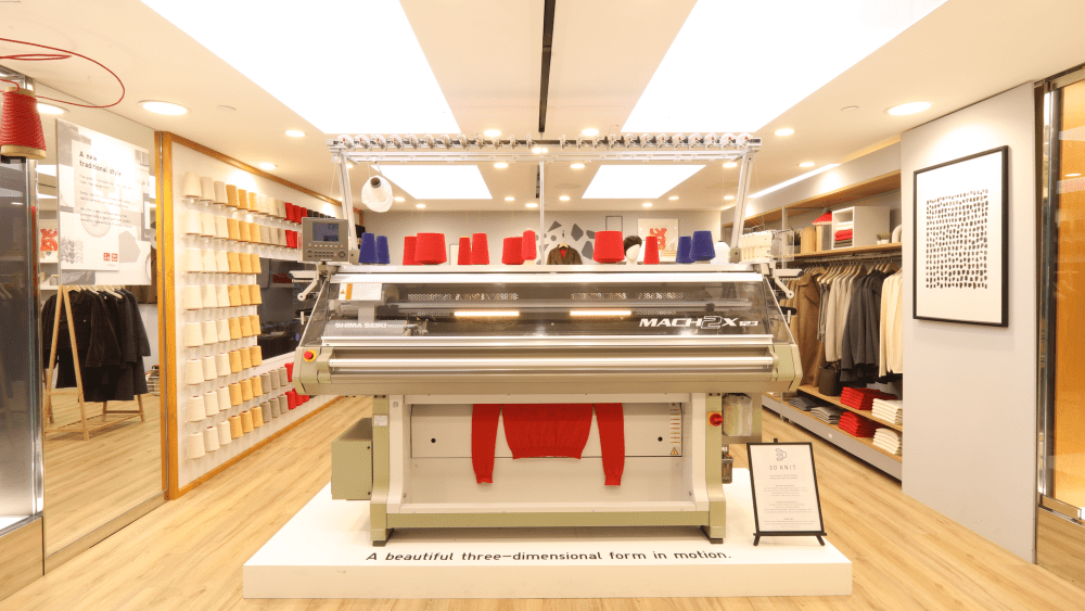 A Uniqlo's 3D knitting machine set up in the fluorescent storefront to artfully knit a red knit sweater.