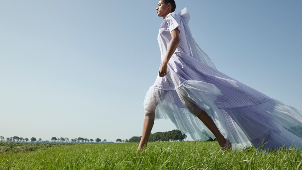 A look from the Viktor & Rolf Tulle collection features a woman walking in a field with blue skies, wearing a long, flowy dress with tulle train trailing behind her.
