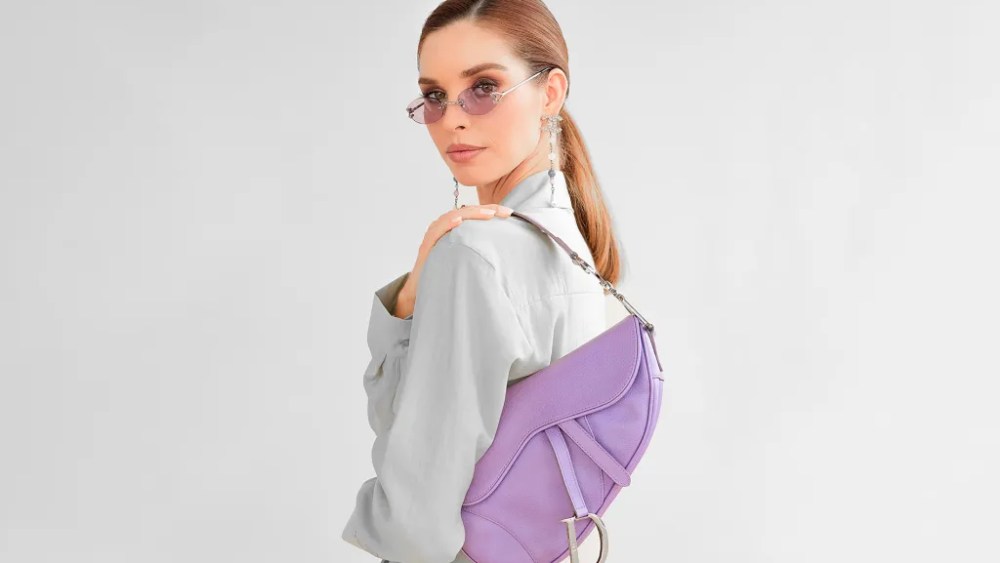 A model in a sleek pony and grey blazer poses in purple sunnies and a purple Dior saddlebag hangs over her shoulder.