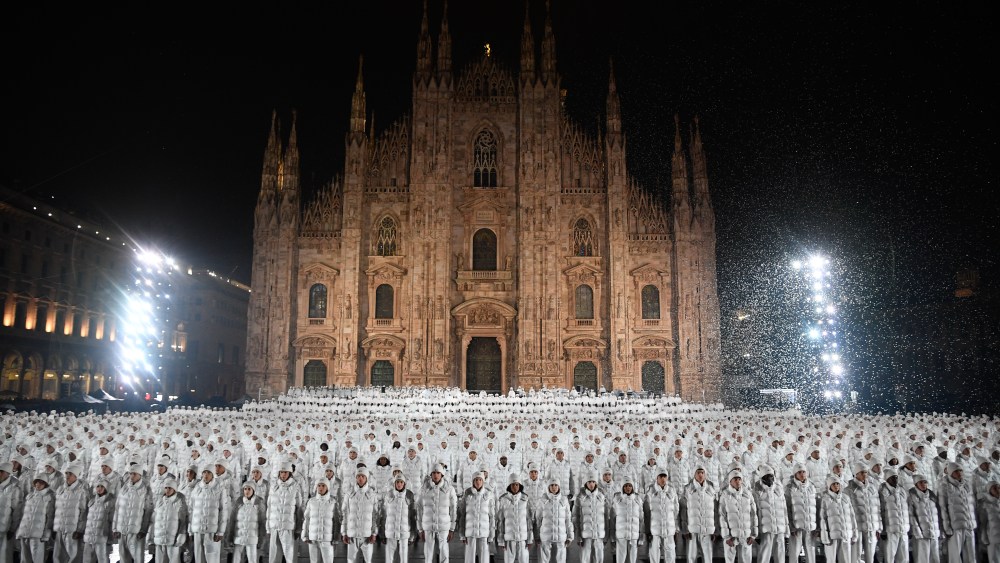 Moncler RTW Spring 2023 features hundreds of people all dressed alike in white Moncler Maya jackets and white pants and shoes, standing in front of Milan's Duomo Cathedral.