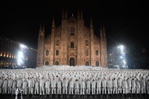 Moncler RTW Spring 2023 features hundreds of people all dressed alike in white Moncler Maya jackets and white pants and shoes, standing in front of Milan's Duomo Cathedral.