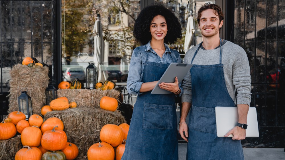 Happy young coffee shop owners standing outdoor near their cafe with laptop and decorating pumpkins preparing for halloween on background.