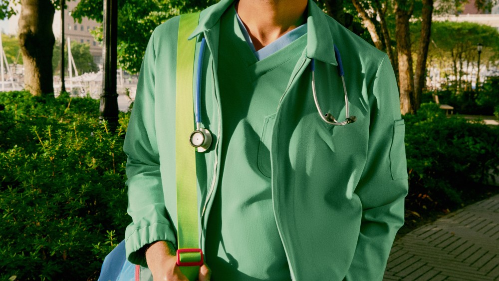 Welles launched as an eco-chic alternative to scrubs. Here, a model sports a green top and jacket with blue pant, striding with a green backdrop.
