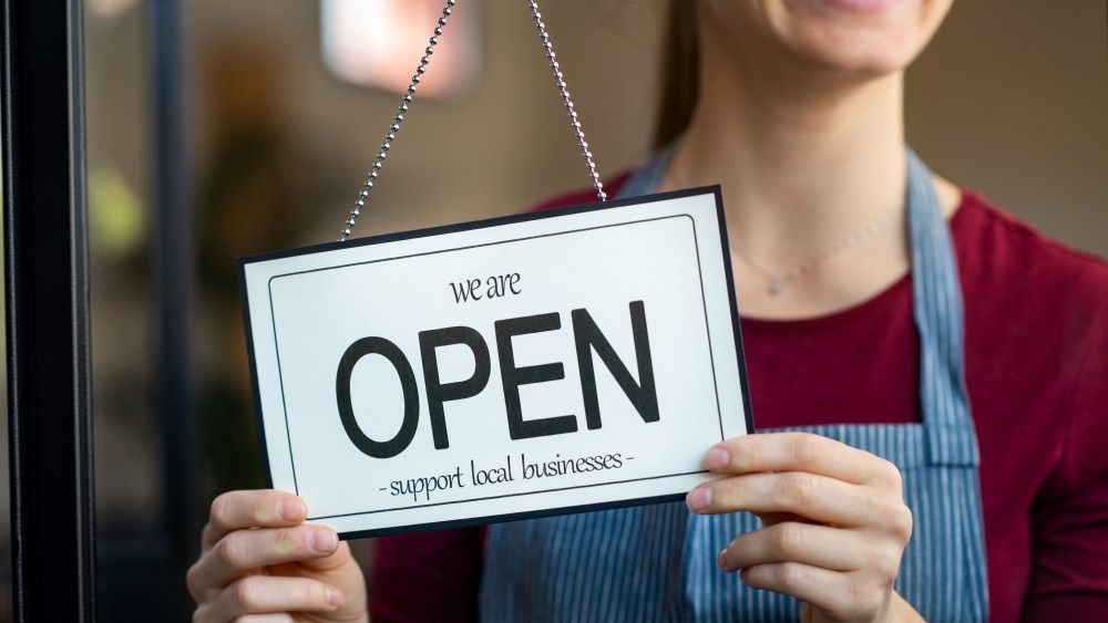 Small business owner smiling while turning the sign for the reopening of the place after the quarantine due to covid-19. Happy businesswoman standing at her restaurant or coffee shop gate with open signboard. Close up of woman’s hands holding sign now we are open support local business.