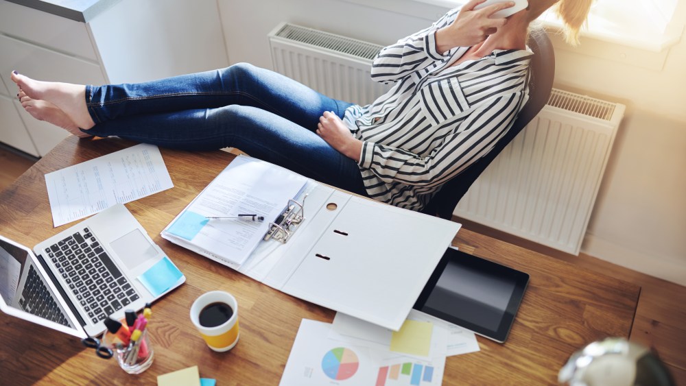 Relaxed confident female entrepreneur reclining back in her chair with her bare feet on the desk talking to a client on her mobile phone, high angle view on desk with paperwork, charts and laptop