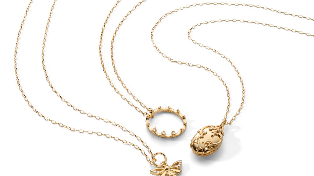The Bridgerton x Monica Rich Kosann collection's core pieces feature a gold bee pendant on a gold chain on the left, a gold poesy ring shaped like a tiara or crown on a gold chain, and a gold locket on a gold chain on the right.