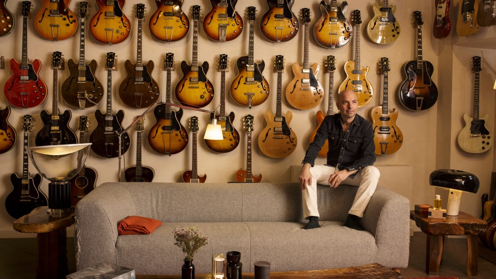 Julian Bedel, founder of Fueguia 1833, at his home in Milan sitting on the headrest of a couch, his feet on the seat, in front of a wall of more than 20 guitars.