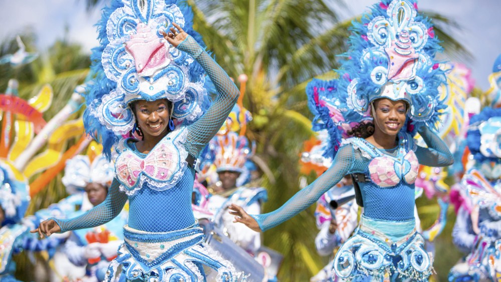 Two women Junkanoo performers in The Bahamas wear blue, white and pink costumes, with cardboard wasitbands, breastplates and large headpieces. They are smiling and appear to be mid-dance.