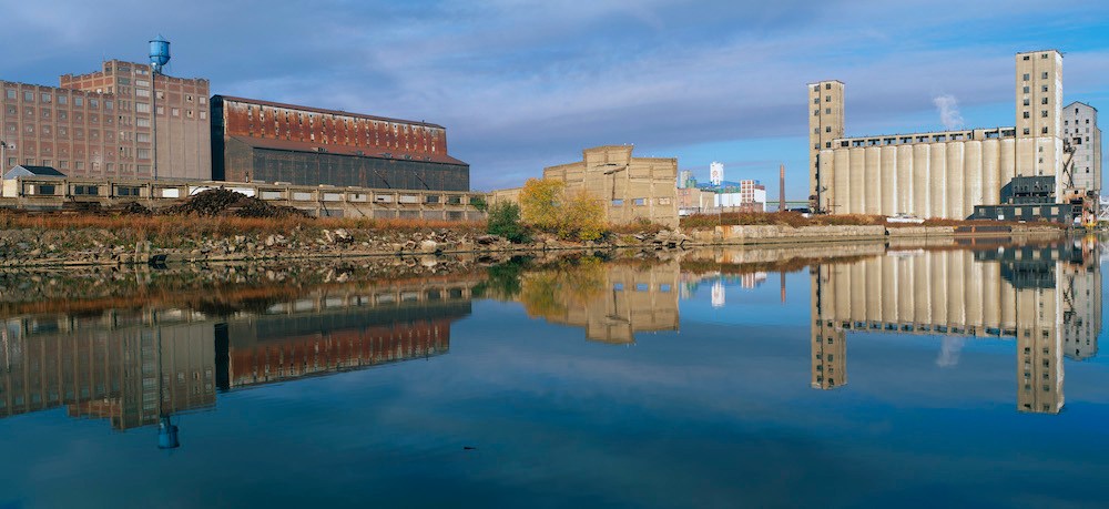 This shows an industrial building through a clearing of trees on the Rogue River in Michigan