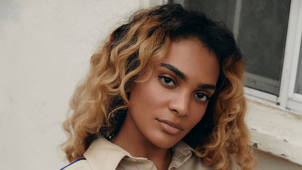 Sydney "Sydeon" Parker sits next to a window wearing a khaki colored collared shirt and white bottoms.