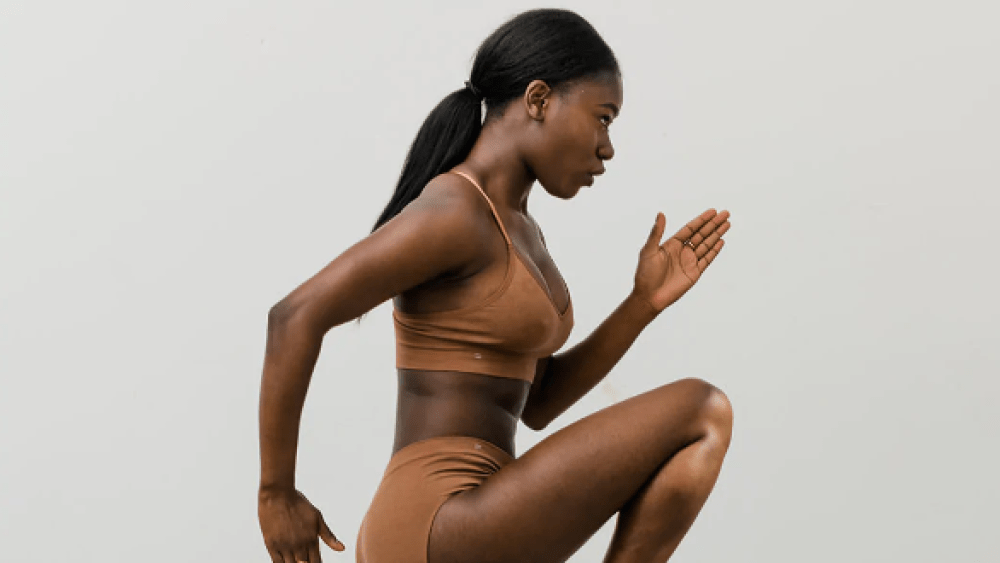 A Black woman wearing nude innerwear from startup Branwyn posing in a jump with arms in blades akin to a runner's warmup.